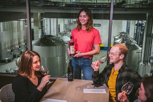 Mari wine server smiling next to guests doing a tasting on the mezzanine level, overlooking the Mari production floor